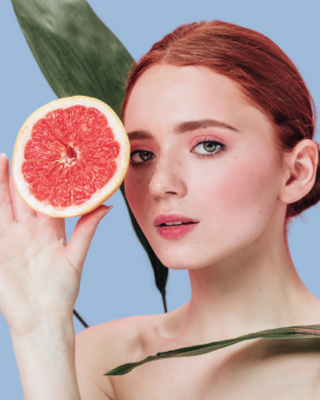 Red-haired woman holding a halved grapefruit to her face with green leaves on a blue background