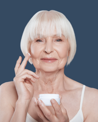 Elderly woman with white hair applying cream to her face on a blue background.