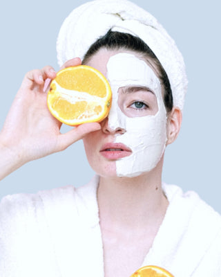 Woman with face mask, holding a halved orange to her face, wearing a white robe, on a blue background